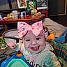 baby, child, infant, smiling, happy, tongue_out, bow, headband, portrait, sitting, activity_seat, toy, colorful, indoor, living_room, blanket, pillow, furniture, raised_fist, close_up