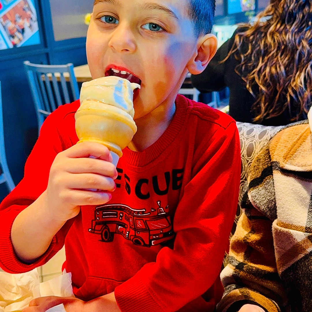 Gio joined the competition — help win amazing prizes! child, boy, ice_cream, cone, eating, smiling, restaurant, indoor, portrait, red_shirt, sweatshirt, fire_truck, table, food, dessert, seated, phone, adult, background, photograph