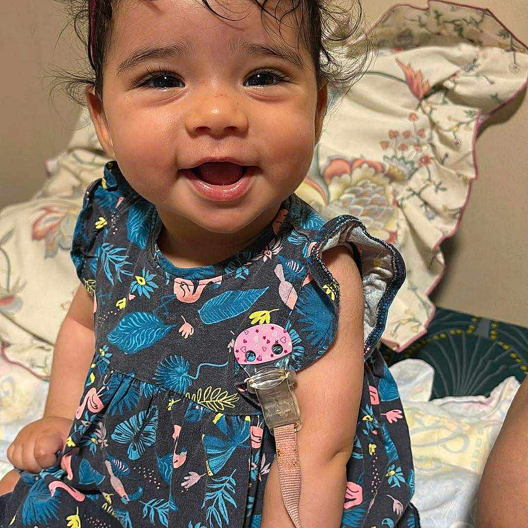 Dalia a rejoint le concours — aidez-le/la à gagner de superbes lots ! accessory, baby, bed, child, comfort, cute, dress, face, flower, hand, happy, headband, indoor, infant, pacifier, patterned_clothing, person, pillow, skin, smiling