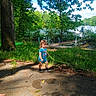 child, toddler, outdoors, lakeside, lake, dock, trees, forest, nature, greenery, path, sidewalk, sunlight, summer, blue_shirt, standing, portrait, water, woodland, family_photo