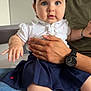 baby, child, man, holding, watch, hand, face, blue_eyes, hair_clip, white_shirt, navy_skirt, jeans, sitting, indoor, person, parent, family, curious, cute, portrait