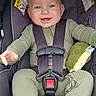 baby, child, car_seat, seatbelt, green_clothing, plush_toy, avocado_toy, smiling, blue_eyes, infant, happy, cute, sitting, child_safety, car_travel, toddler, face, person, indoor, closeup