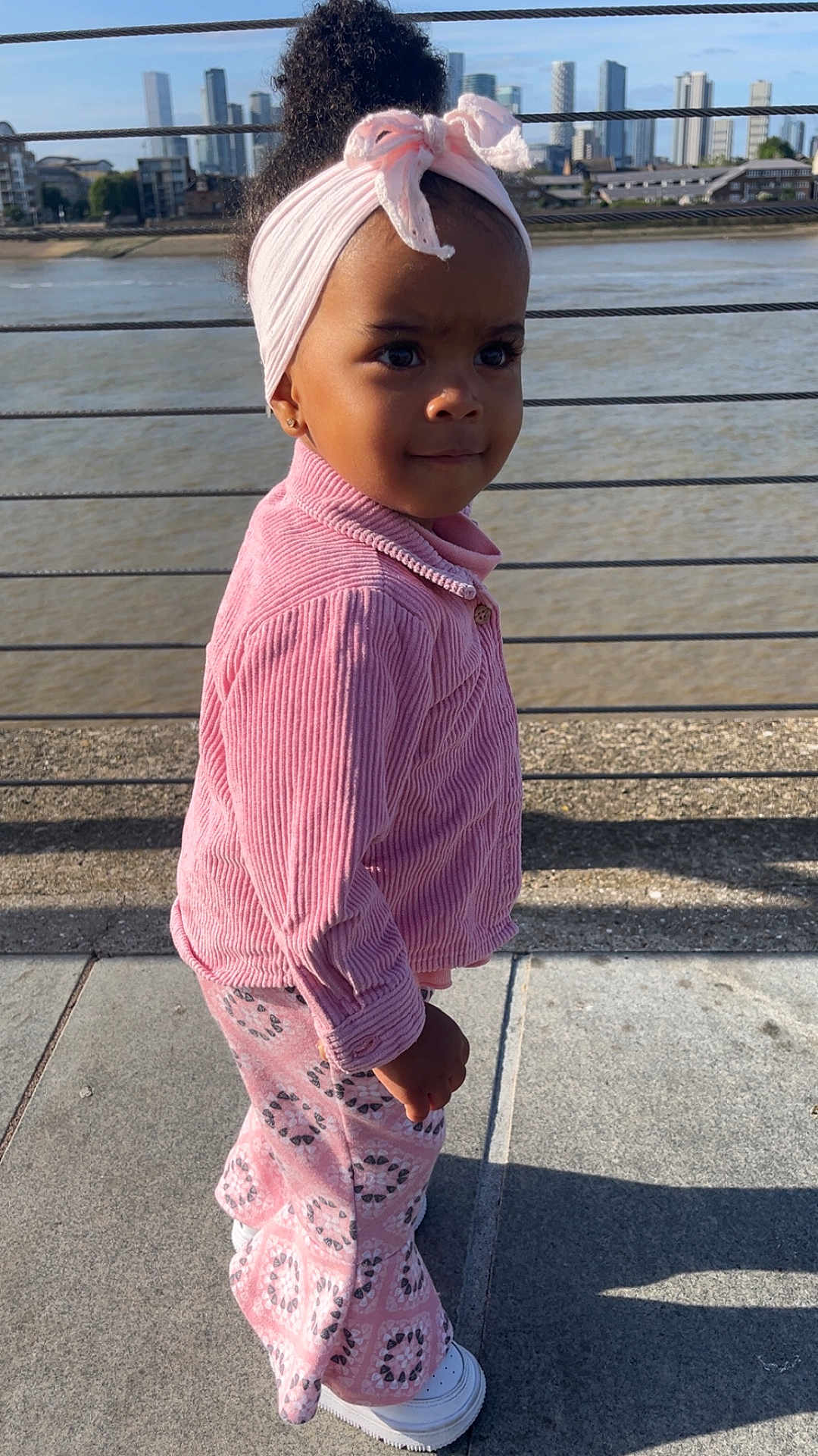 Maria joined the competition — help win amazing prizes! child, toddler, pink_clothing, headband, outdoor, river, cityscape, sidewalk, shadow, building, water, daylight, fashion, portrait, young_child, casual_wear, footwear, urban, smiling, person