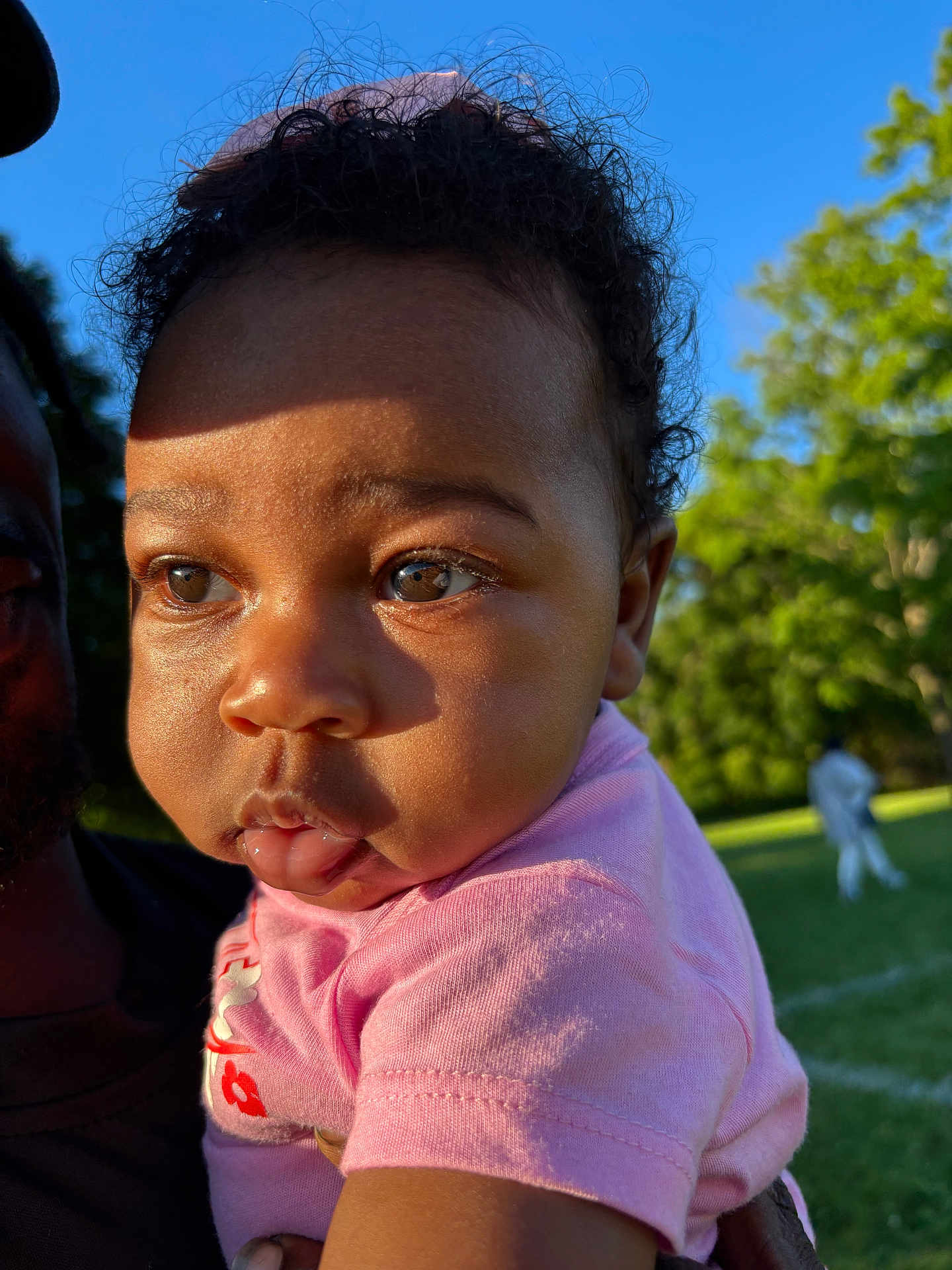 Jordyn is registered to the contest to win money with this photo: baby, child, face, person, outdoor, sunlight, shadow, grass, tree, greenery, park, curly_hair, pink_shirt, expression, portrait, holding, nature, daylight, skin, eyes