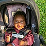 baby, car_seat, child, grass, outdoor, sunlight, smile, curly_hair, bow, clothing, safety_harness, infant, nature, greenery, flowers, person, seat, cute, happy, portrait