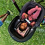baby, stroller, infant, pink_socks, pink_clothing, grass, outdoor, child, legs, person, pavement, carrying_handle, seat_belt, toy, cute, pouting, relaxed, park, daylight, casual