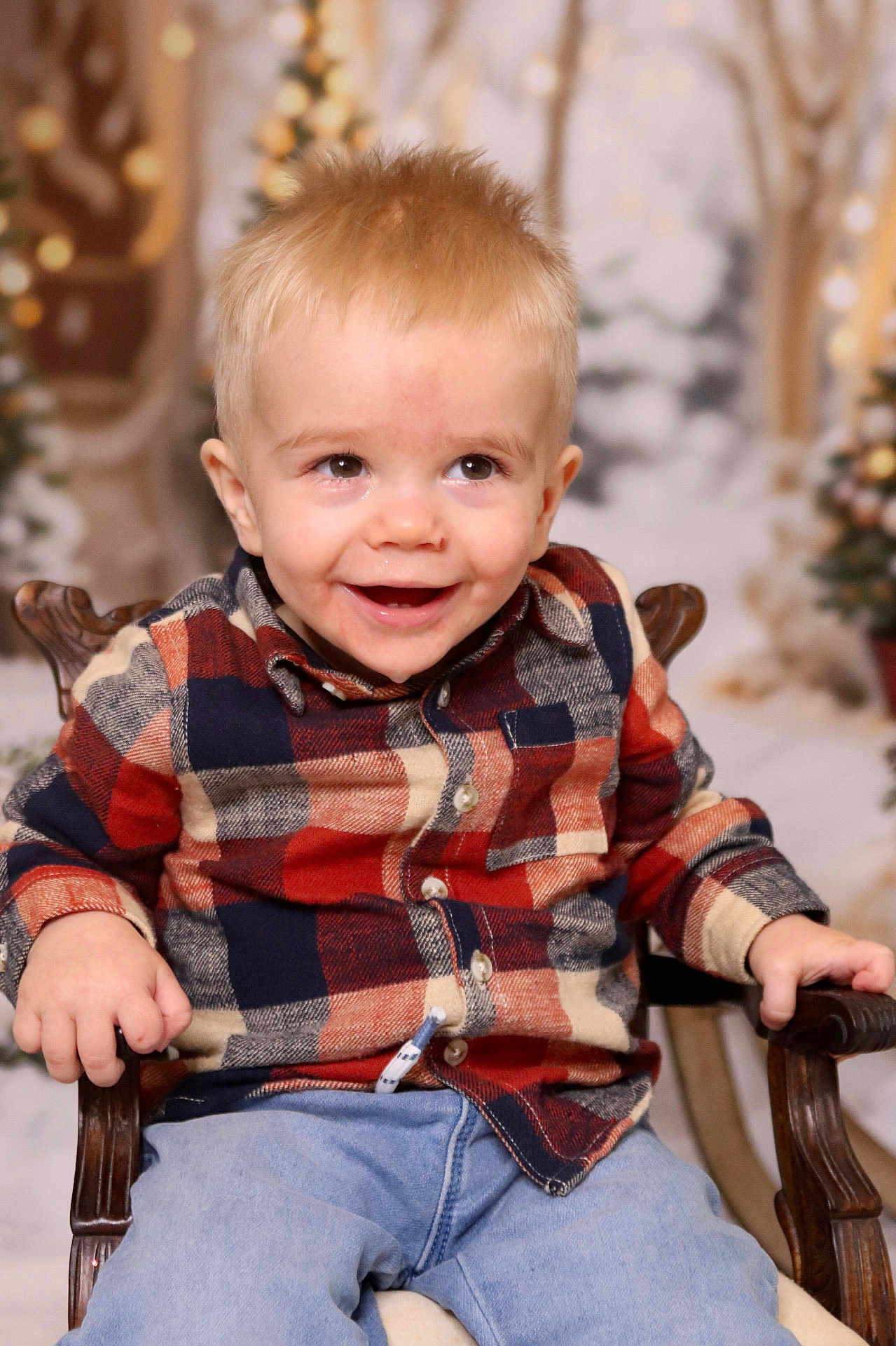Hudson is registered to the contest to win money with this photo: blonde_hair, blue_jeans, casual_clothing, chair, child, cute, face, festive, happy, holiday_background, indoor, person, plaid_shirt, portrait, seated, smiling, toddler, warm_lighting, wooden_chair, young_child