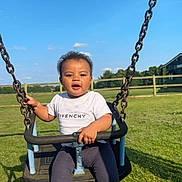 Kaylum is registered to the contest to win money with this photo: toddler, child, swing, playground, outdoor, grass, blue_sky, clouds, sunny, givenchy, white_tshirt, mismatched_socks, happy, holding, metal_chains, fence, park, casual_clothing, daylight, smiling