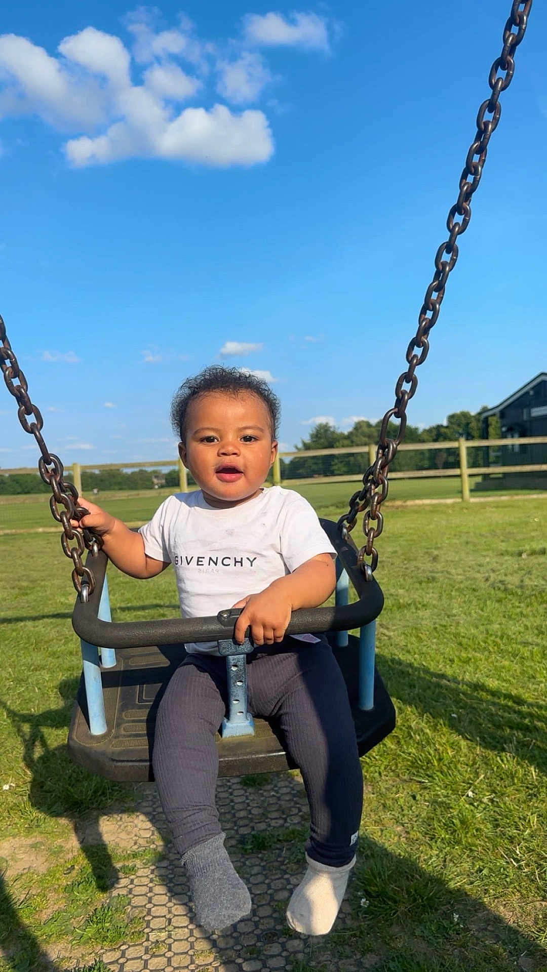 Kaylum is registered to the contest to win money with this photo: toddler, child, swing, playground, outdoor, grass, blue_sky, clouds, sunny, givenchy, white_tshirt, mismatched_socks, happy, holding, metal_chains, fence, park, casual_clothing, daylight, smiling
