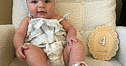 Millie is registered to the contest to win money with this photo: baby, child, infant, headband, bow, chair, plush, soft_texture, wooden_plaque, months_marker, white_sandals, smiling, cute, comfortable, indoors, portrait, happy, sitting, clothing, person