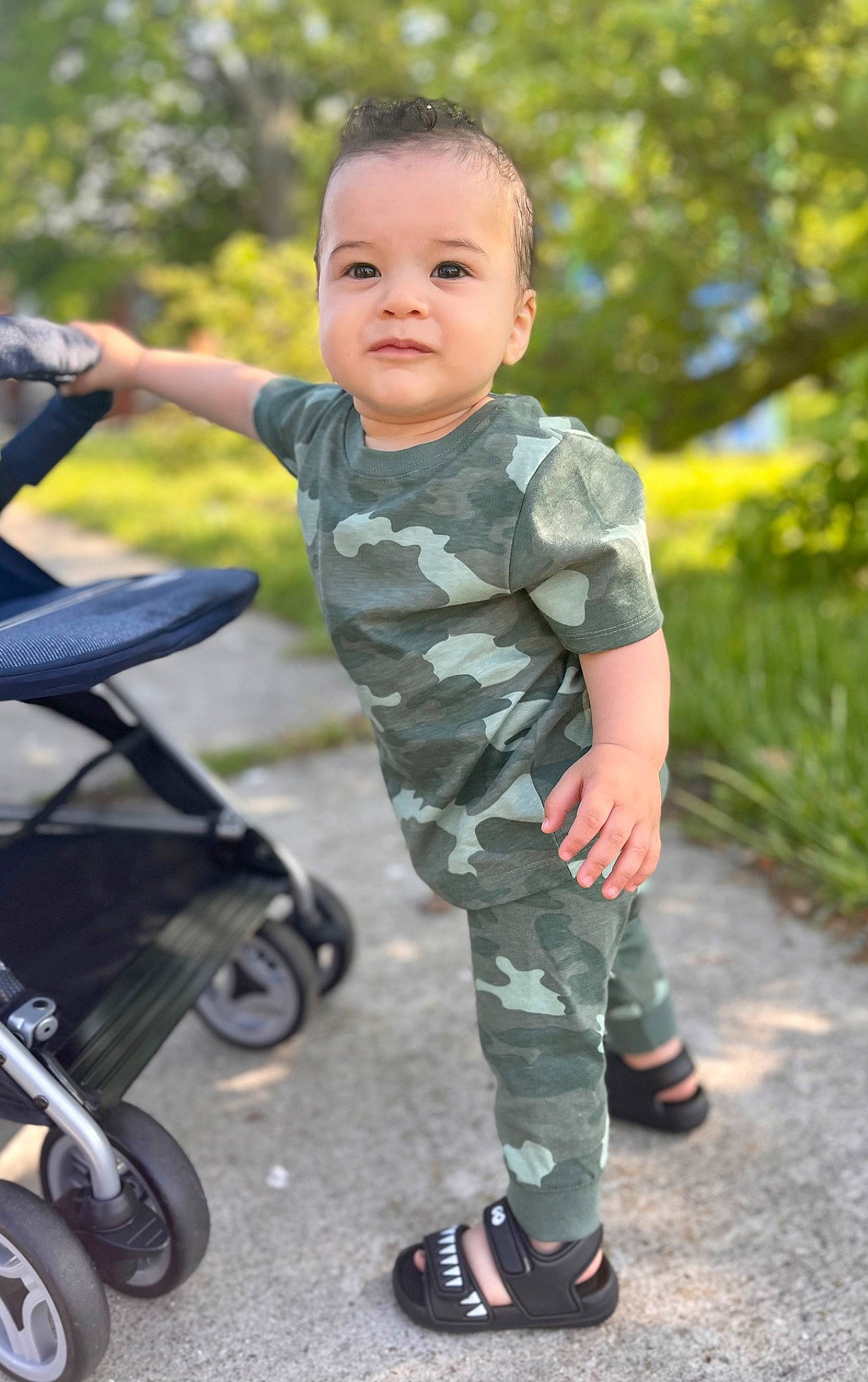 Matthew is registered to the contest to win money with this photo: asphalt, baby, baby_toddler_clothing, camouflage, fun, grass, happy, leisure, military_camouflage, people_in_nature, person, plant, recreation, rolling, sleeve, smile, sneakers, tire, toddler, tree