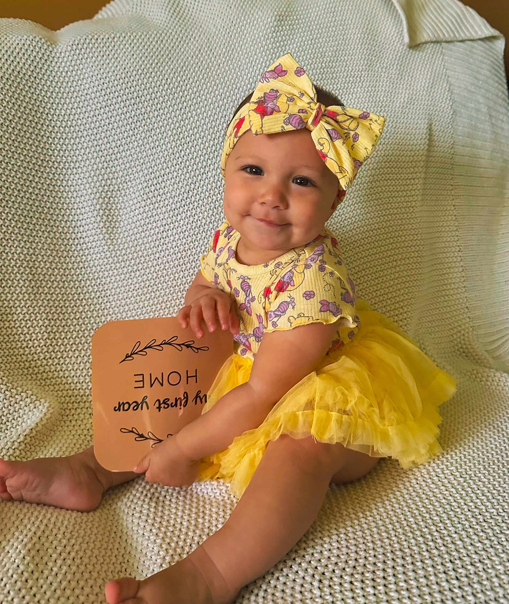 Odayah joined the competition — help win amazing prizes! baby, child, smiling, yellow, headband, dress, sitting, blanket, cute, portrait, happy, infant, clothing, person, indoors, sign, hand, feet, knit, tutu