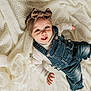 baby, child, infant, denim_overalls, headband, bow, smiling, happy, lying_down, blanket, white_blanket, cute, clothing, portrait, one_person, indoors, baby_hand, baby_face, soft_texture, cozy