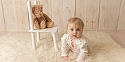 Maddison joined the competition — help win amazing prizes! baby, infant, crawling, white_clothing, plush_toy, teddy_bear, chair, fluffy_rug, wooden_wall, indoors, child, cute, portrait, soft_lighting, cozy, floor, expression, young_child, looking_up, person