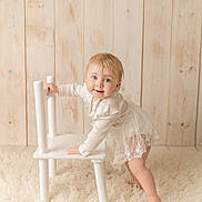 Maddison is registered to the contest to win money with this photo: toddler, child, white_dress, barefoot, chair, fluffy_rug, wooden_wall, indoor, smiling, playful, baby, portrait, cute, soft_lighting, floor, standing, person, young_child, happy, cozy