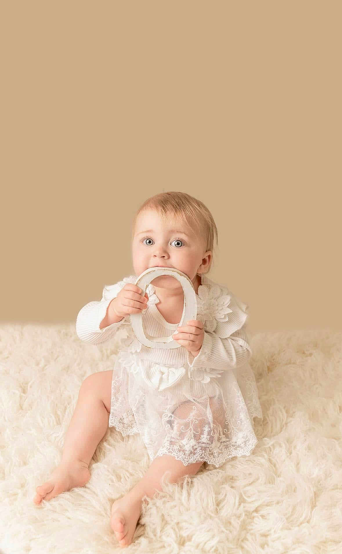 Maddison joined the competition — help win amazing prizes! baby, child, infant, white_dress, lace, fluffy_rug, sitting, holding, letter_o, nibbling, cute, soft_lighting, beige_background, portrait, indoor, happy, young_child, person, smiling, cozy