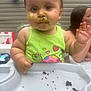 Monroe is registered to the contest to win money with this photo: baby, child, high_chair, food, messy, face_paint, green_clothing, turtle_design, hand, smudged_food, woman, balloon, background, outdoor, table, plate, celebration, curious_expression, person, portrait