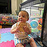 baby, child, indoor, playpen, toys, colorful, foam_mats, smiling, wooden_plaque, storage_bin, window, daylight, happy, sitting, clothing, shorts, tshirt, floor, playroom, person