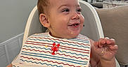 Jt is registered to the contest to win money with this photo: baby, high_chair, bib, food, scrambled_eggs, smile, happy, face, hand, indoor, home, kitchen, child, person, furniture, plate, table, clothing, feeding, messy