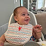 baby, high_chair, bib, food, scrambled_eggs, smile, happy, face, hand, indoor, home, kitchen, child, person, furniture, plate, table, clothing, feeding, messy