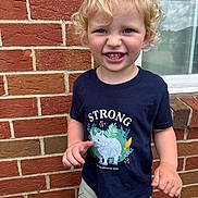 Everett is registered to the contest to win money with this photo: toddler, child, smiling, curly_hair, blonde_hair, navy_shirt, rhino_graphic, text_on_clothing, brick_wall, window, outdoor, green_shorts, happy, portrait, person, young_child, cute, daylight, casual_clothing, nature