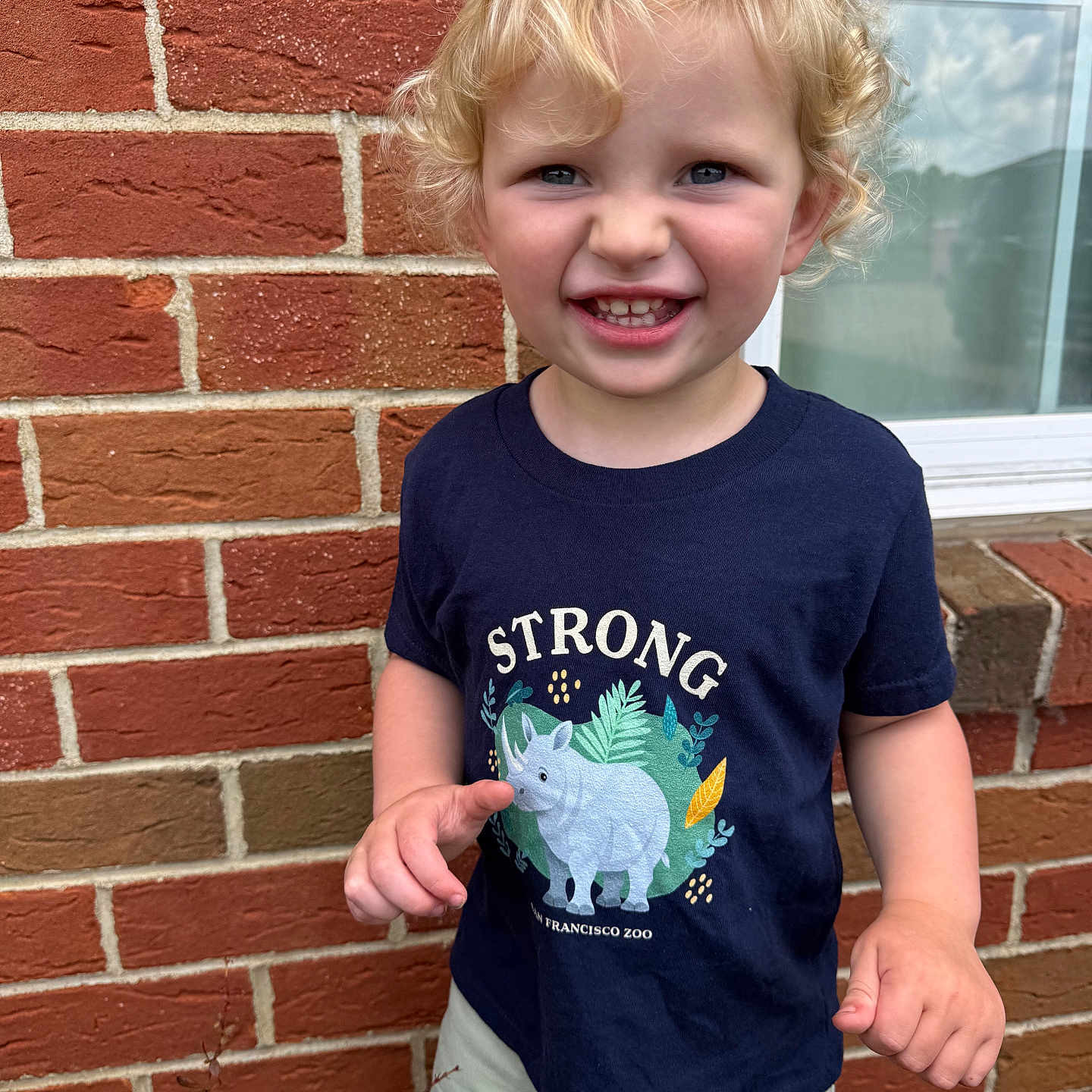 Everett is registered to the contest to win money with this photo: toddler, child, smiling, curly_hair, blonde_hair, navy_shirt, rhino_graphic, text_on_clothing, brick_wall, window, outdoor, green_shorts, happy, portrait, person, young_child, cute, daylight, casual_clothing, nature