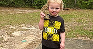Everett is registered to the contest to win money with this photo: toddler, child, smiling, outdoor, forest, trees, grass, concrete, nirvana_tshirt, black_clothing, shorts, crocs, holding_object, rock, nature, young_child, standing, daylight, curly_hair, playful