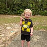 toddler, child, smiling, outdoor, forest, trees, grass, concrete, nirvana_tshirt, black_clothing, shorts, crocs, holding_object, rock, nature, young_child, standing, daylight, curly_hair, playful