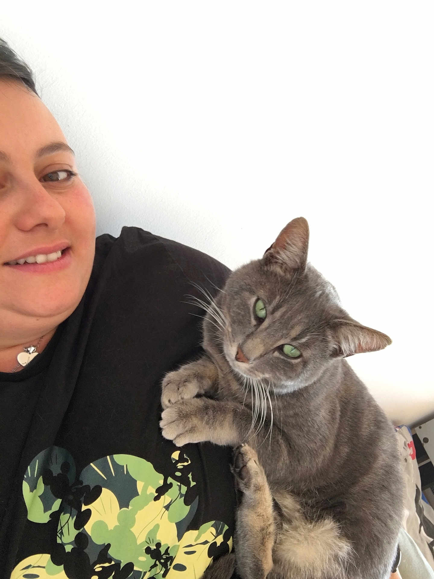 Oziris participe au concours pour gagner de l'argent avec cette photo : person, cat, smiling, cuddling, gray_cat, green_eyes, indoor, casual, portrait, close_up, black_shirt, mickey_mouse_print, short_hair, light_skin, white_background, pet, friendly, relaxed, feline, happy