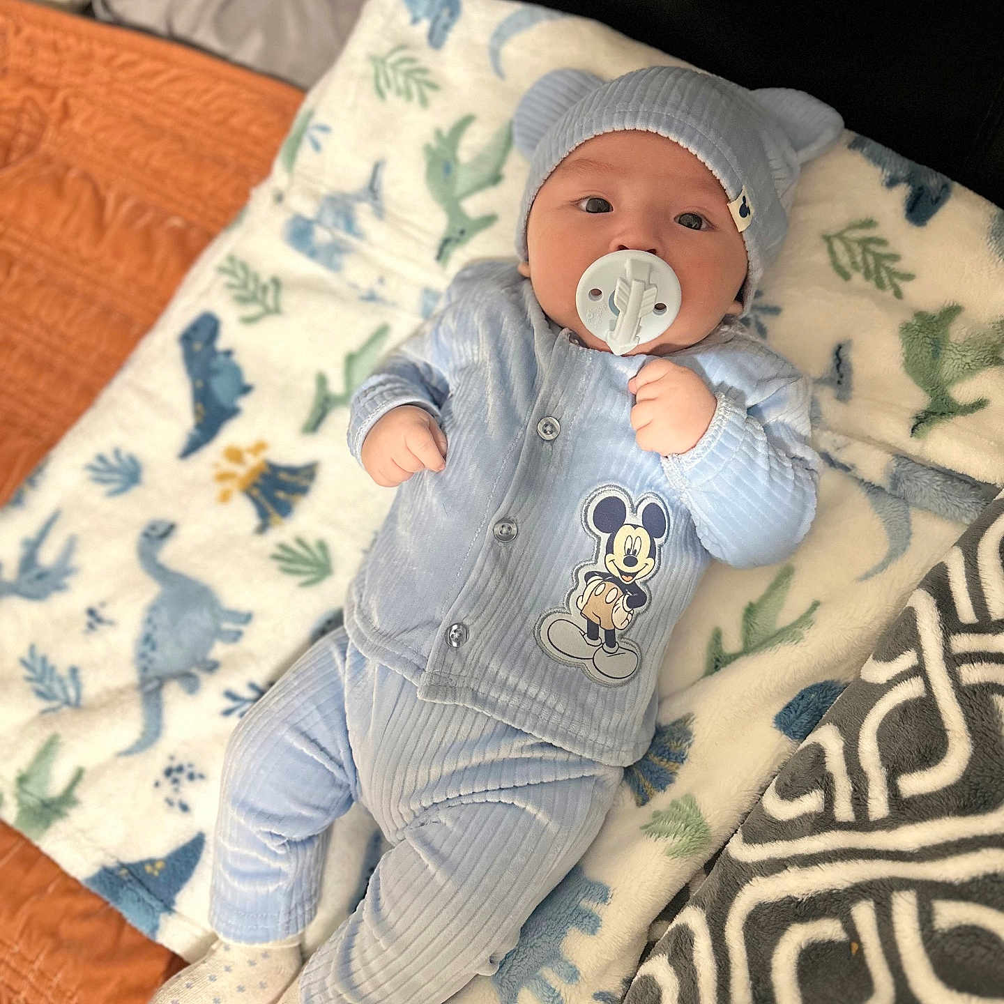 Sasha is registered to the contest to win money with this photo: baby, baby_boy, bear_ears, blanket, blue_outfit, child, comfortable, cute, dinosaur_pattern, hat, headwear, indoor, infant, lying_down, mickey_mouse, pacifier, portrait, sleepwear, socks, soft_texture
