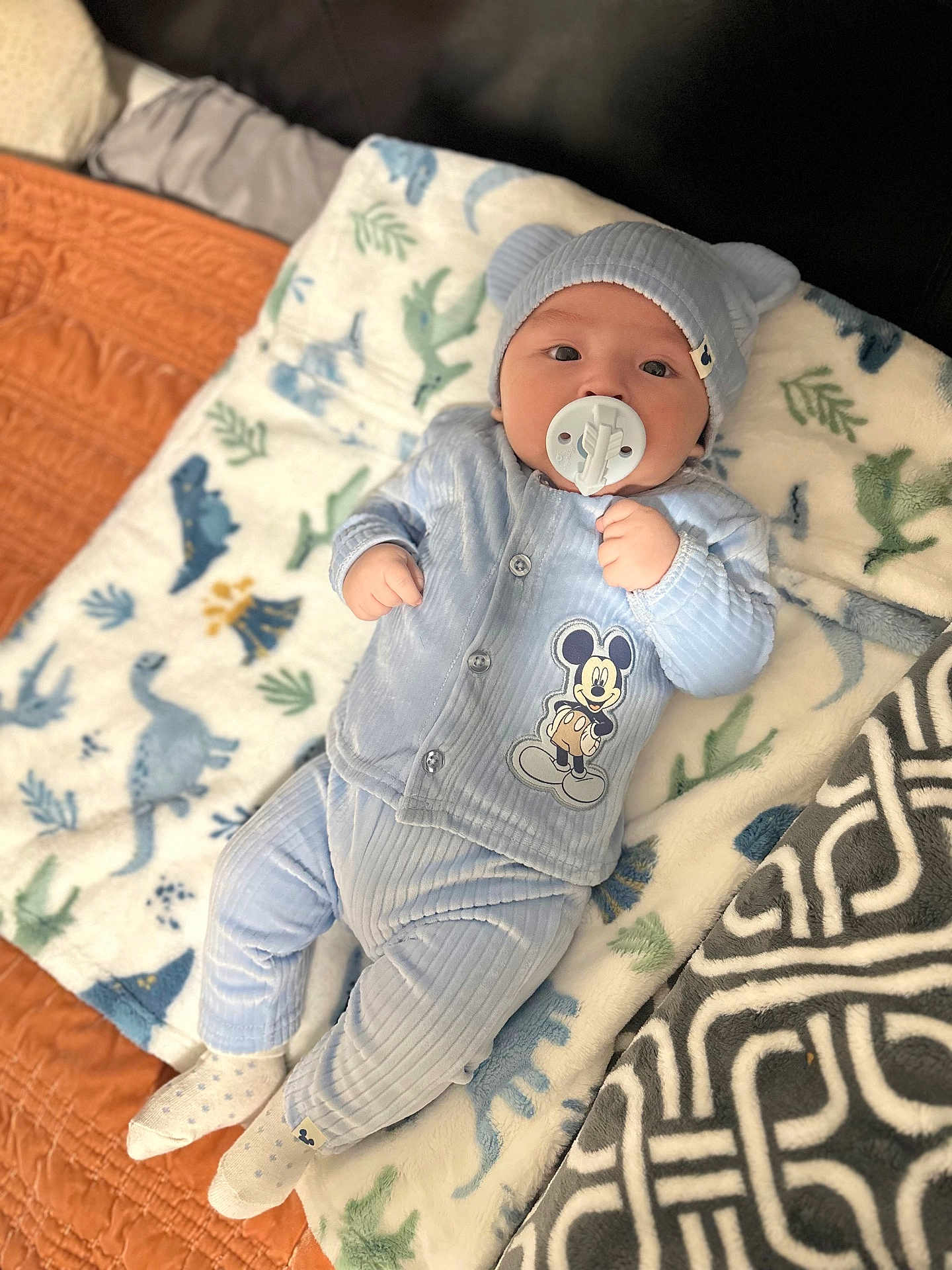 Sasha is registered to the contest to win money with this photo: baby, pacifier, blue_outfit, mickey_mouse, hat, bear_ears, blanket, dinosaur_pattern, socks, infant, cute, lying_down, soft_texture, indoor, child, portrait, comfortable, sleepwear, headwear, baby_boy