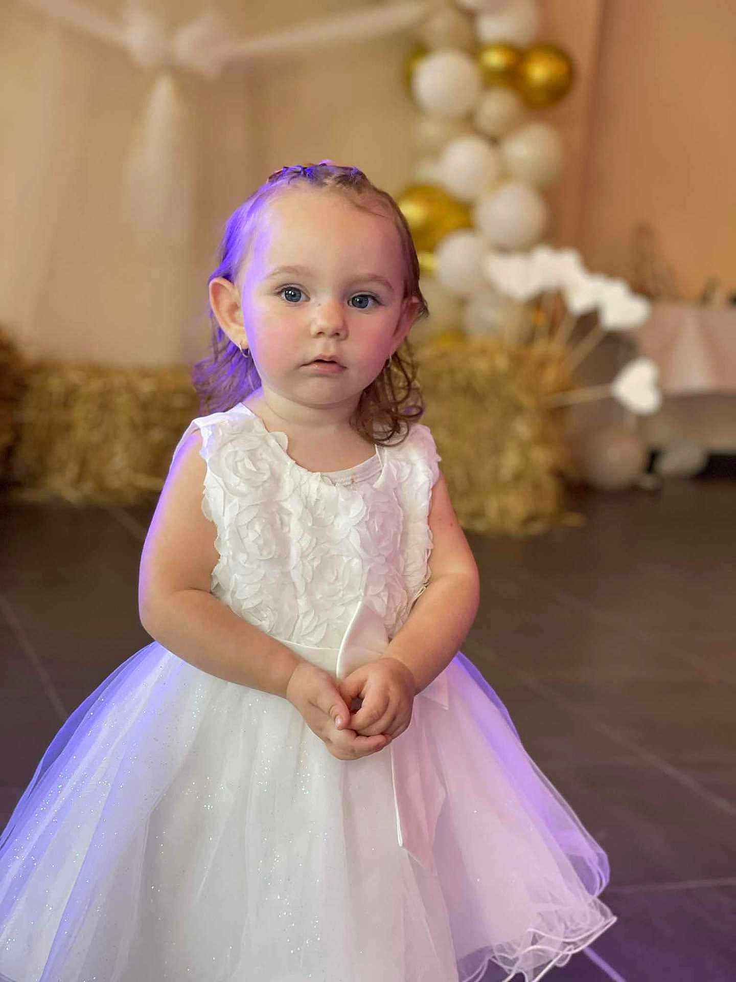 Kamèlya a rejoint le concours — aidez-le/la à gagner de superbes lots ! child, girl, white_dress, portrait, indoor, balloons, decoration, curious_expression, hands_clasped, young_child, event, celebration, floor_tiles, soft_lighting, face, ears, blue_eyes, short_hair, tulle_skirt, bow