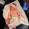 baby, infant, blanket, pink_clothing, headband, pacifier, smartphone, adult_hand, jeans, patterned_blanket, newborn, lying_down, cute, indoor, soft_lighting, small_feet, person, clothing, accessory, resting