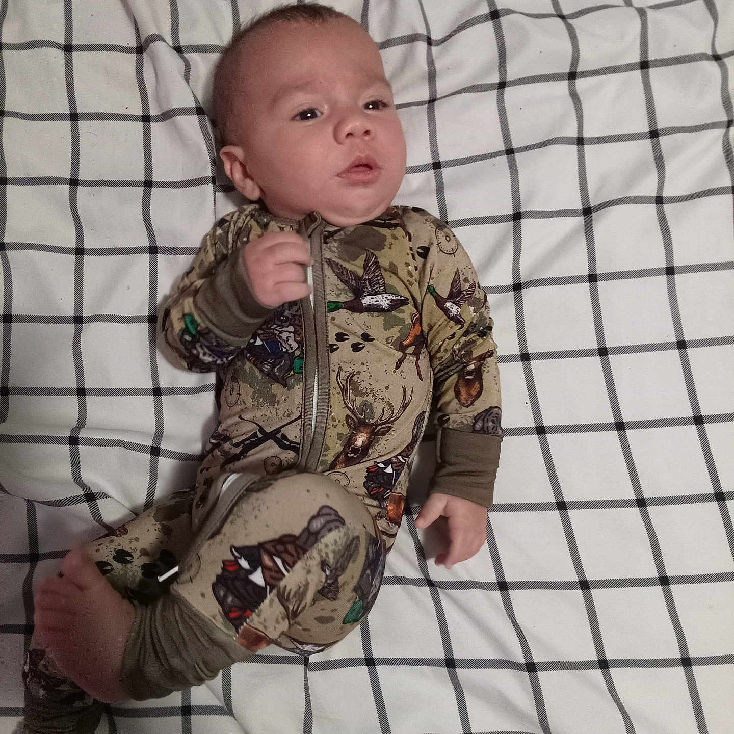 Bruce is registered to the contest to win money with this photo: baby, bed, blanket, camo_pattern, checkered, clothing, curious, expression, face, foot, hand, head, indoor, infant, lying_down, onesie, person, portrait, sleepwear, young_child