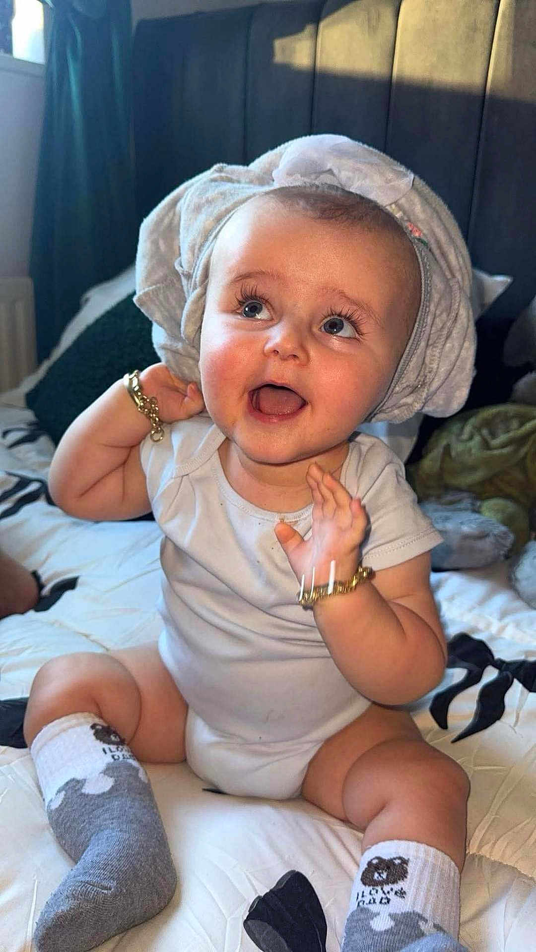 Freddie is registered to the contest to win money with this photo: baby, child, smiling, sitting, bed, towel, onesie, socks, bracelet, happy, indoors, cute, portrait, infant, headwear, expression, skin, person, face, candid