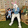child, bunny_costume, person, bench, paper_chains, decorations, carrots, flowers, grass, indoor, smiling, blue_clothing, toy, holiday_theme, celebration, happy, costume, seated, playful, event