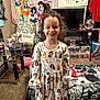 child, smiling, curly_hair, dress, princess_characters, doll, bedspread, spider_web_pattern, pop_culture_collectibles, chair, pillow, television, carpet, table, figurines, shelf, room, indoor, happy, person