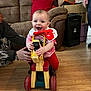 baby, child, smiling, toy, ride_on_toy, wooden_floor, living_room, couch, bib, red_clothing, boots, adult_hand, cardboard_box, smile, furniture, person, happy, play, indoor, flooring