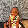 toddler, child, smiling, colorful_vest, white_dress, sitting, indoor, floor, happy, cute, portrait, face, person, earrings, texture, couch, wooden_floor, bright_eyes, joyful, young_child