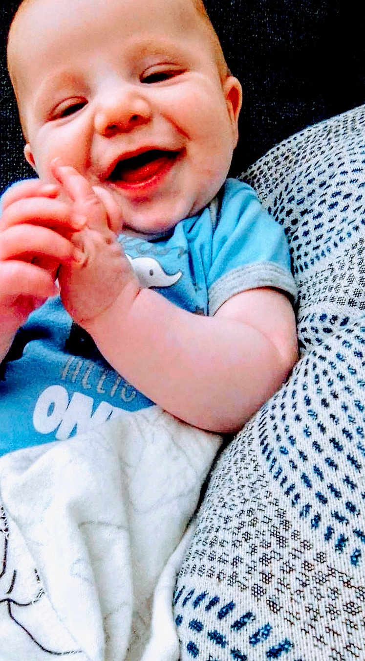 Josiah joined the competition — help win amazing prizes! baby, smiling, blue_shirt, hands, blanket, cushion, patterned, infant, happy, close_up, face, child, cute, resting, indoor, person, soft, cozy, playful, young