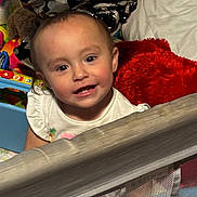 Skylar is registered to the contest to win money with this photo: toddler, child, smile, face, playpen, toy, pillow, red_pillow, colorful, indoor, baby, clothing, white_shirt, mesh, happy, person, head, cute, young_child, furniture