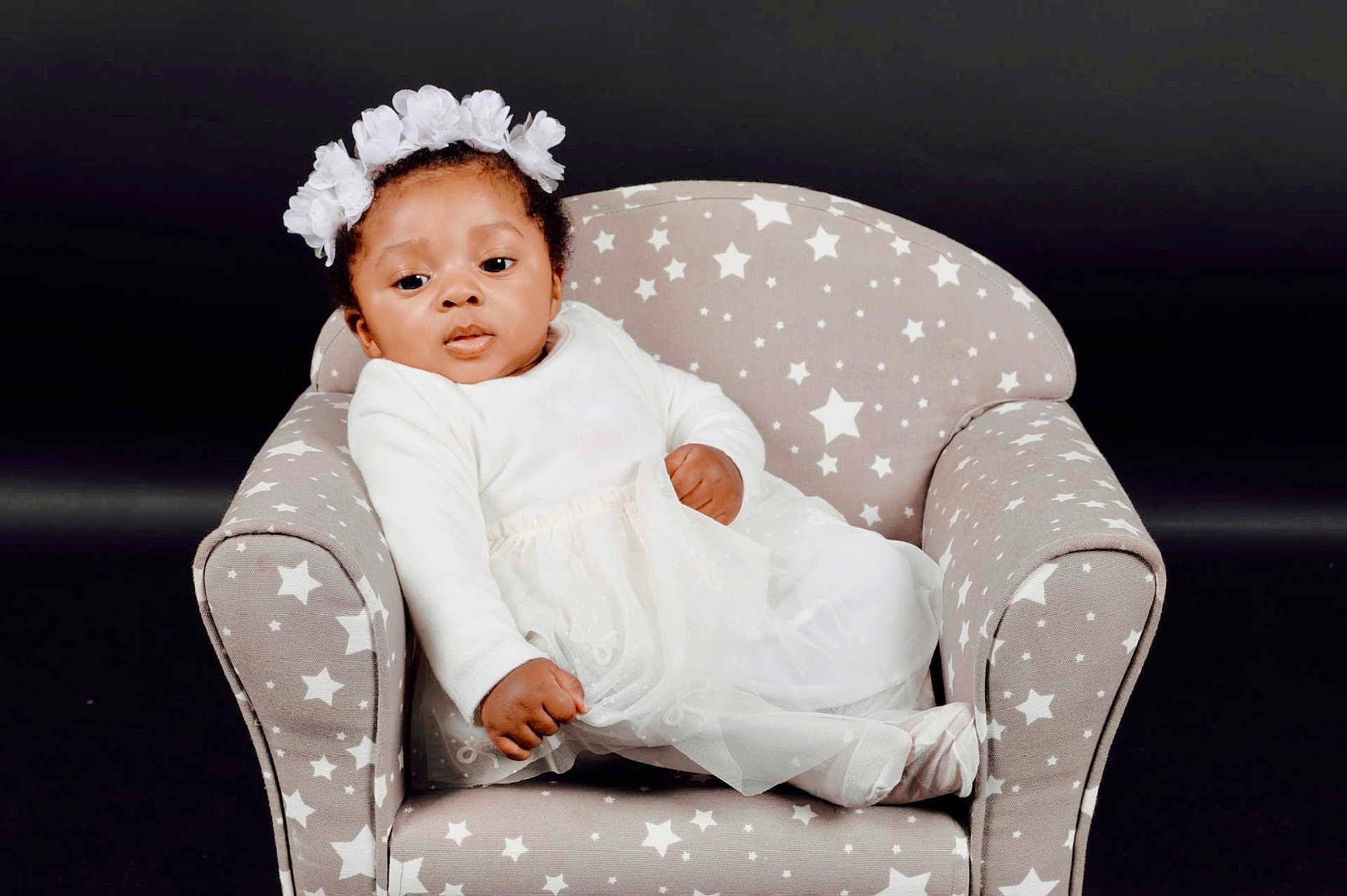 Norah participe au concours pour gagner de l'argent avec cette photo : baby, child, infant, white_clothing, headband, flower_crown, armchair, star_pattern, tulle_skirt, portrait, studio_photo, dark_background, cute, relaxed, seated, soft_fabric, indoor, one_person, young_child, fashion