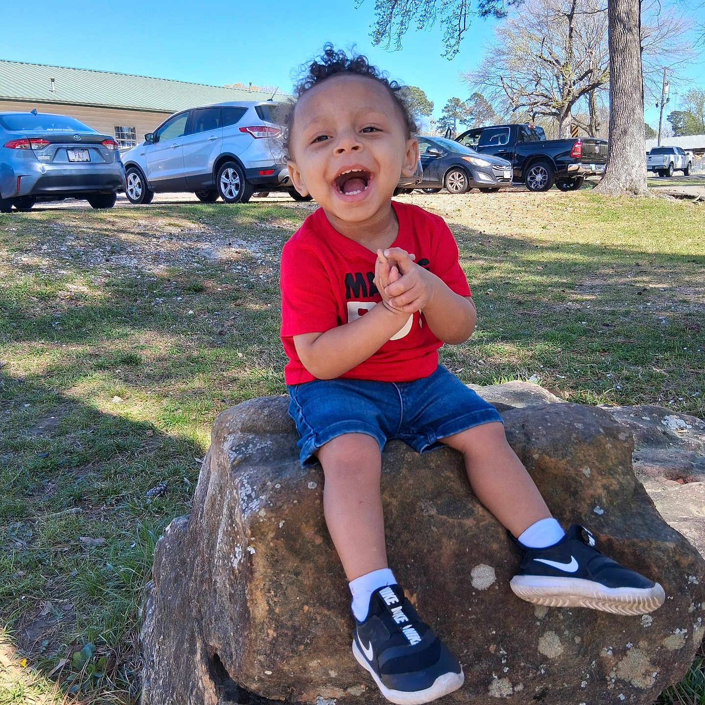 Xavier is registered to the contest to win money with this photo: toddler, child, rock, grass, tree, outdoor, blue_sky, car, laughing, clapping, curly_hair, red_shirt, blue_shorts, nike_shoes, white_socks, happy, sunny, parked_cars, nature, daylight