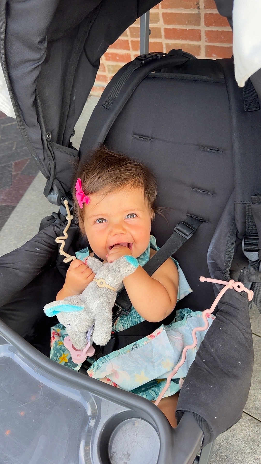 Maleia is registered to the contest to win money with this photo: baby, child, stroller, toy, stuffed_animal, pink_bow, smiling, happy, cute, infant, seatbelt, outdoor, dress, person, face, hands, playful, cheerful, bright, daylight