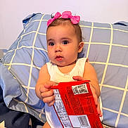 Athena joined the competition — help win amazing prizes! baby, child, pink_bows, pillow, snack_package, white_onesie, indoor, curious, sitting, holding, face, headwear, cute, expression, person, furniture, home, young_child, portrait, bright_eyes
