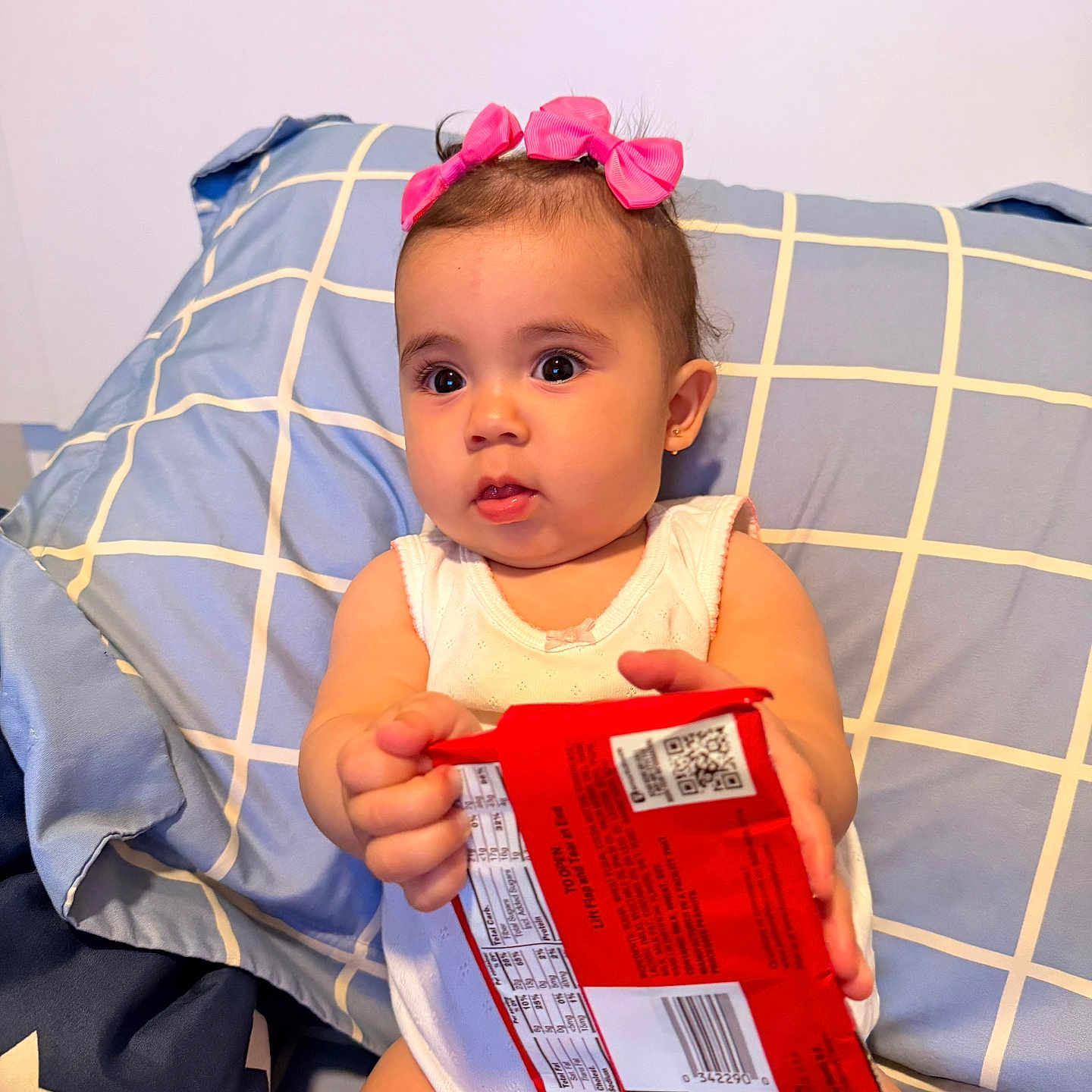 Athena joined the competition — help win amazing prizes! baby, child, pink_bows, pillow, snack_package, white_onesie, indoor, curious, sitting, holding, face, headwear, cute, expression, person, furniture, home, young_child, portrait, bright_eyes
