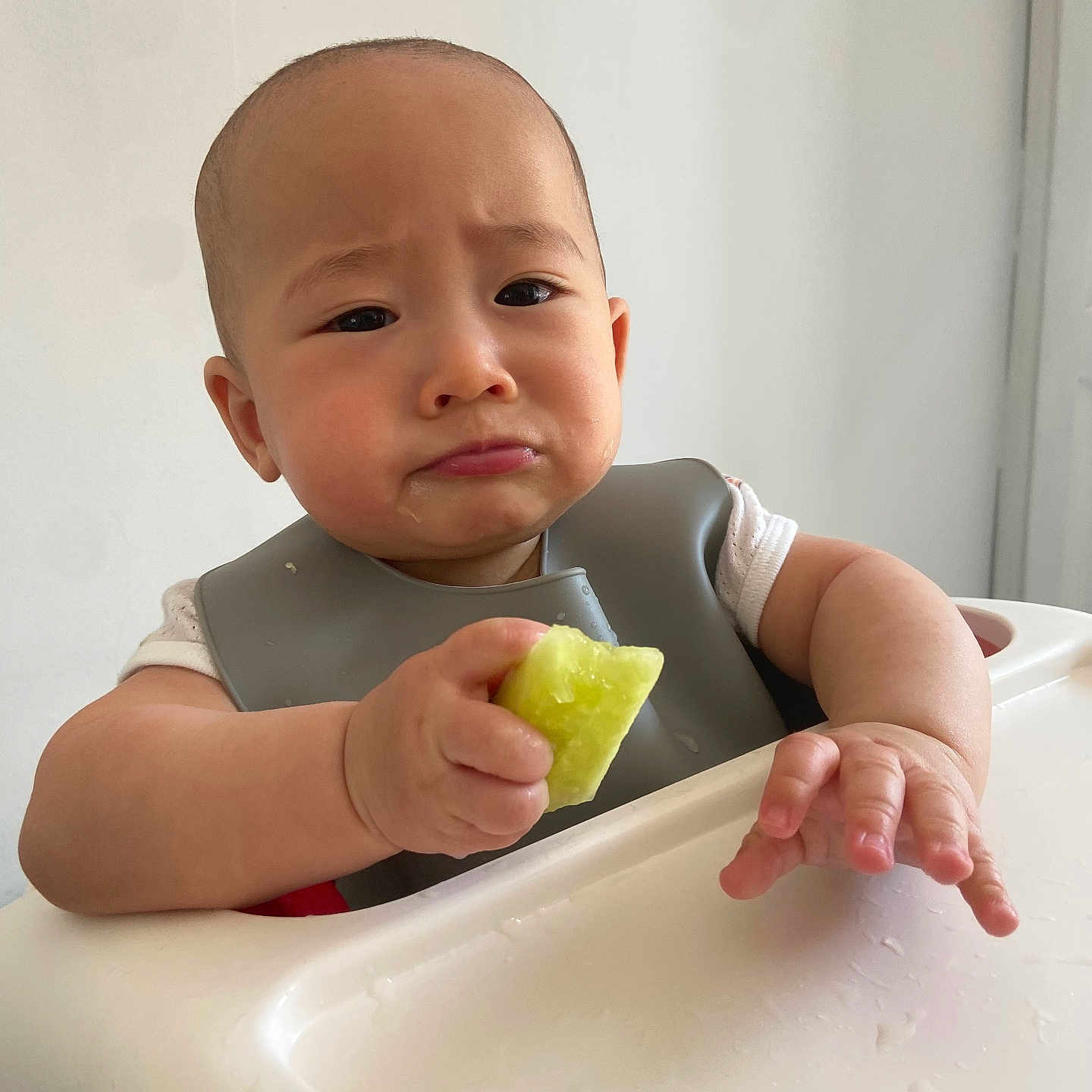 Paxton is registered to the contest to win money with this photo: baby, bib, child, clothing, cute, expression, face, food, fruit, green, hand, high_chair, indoor, person, portrait, sitting, skin, table, white_wall, young