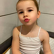 Lina a rejoint le concours — aidez-le/la à gagner de superbes lots ! child, girl, white_tank_top, gray_pants, puckered_lips, sitting, cross_legs, indoor, tiled_wall, playful_expression, face, arms, casual_clothing, light_skin, young, portrait, cute, person, head, hair