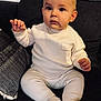 baby, child, infant, sitting, white_clothing, knitwear, couch, pillow, indoor, cute, face, expression, hands, footwear, soft_lighting, portrait, young_child, cozy, curious, casual