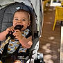 baby, stroller, remote_control, smiling, child, infant, seat, harness, clothing, striped, bear_print, indoor, wooden_floor, yellow_chair, blurred_background, happy, chewing, playful, person, casual